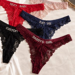 Peut inclure: Un ensemble de cinq strings en dentelle rouge, rose, bleu marine, noir et bordeaux. Chaque pièce présente des lettres en strass sur la ceinture, affichant les inscriptions "SEXY BABY", "EAT ME", "CUSTOM" et "DADDY'S GIRL" en cristaux argentés.