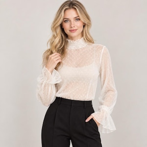 Puede incluir: Una blusa de color crema de manga larga con una capa de malla transparente de lunares, cuello alto fruncido y puños con volantes. La prenda se lleva sobre una camiseta interior de color crema a juego, combinada con pantalones negros.