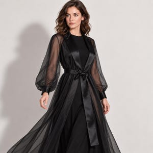 Könnte beinhalten: Ein bodenlanges schwarzes Kleid mit einem blickdichten Oberteil und einem transparenten Tüllüberwurf. Das Kleid hat lange, voluminöse Bischofsärmel mit Satinbündchen, einen Satin-Reverskragen und einen passenden Bindegürtel aus Satin.