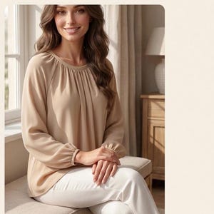 Puede incluir: Una blusa beige de manga larga con cuello redondo y detalles de tela fruncida en el cuello y los puños. El texto GATHERED BLOUSE aparece en la parte superior de la imagen. La prenda se combina con pantalones blancos.