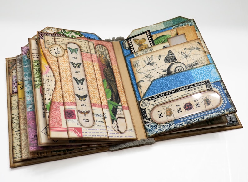 Op de afbeelding: Een handgemaakt scrapbook met een blauwe en witte bedrukte omslag. Het scrapbook is open om verschillende pagina's te laten zien met verschillende patronen en afbeeldingen van insecten, vlinders en bloemen. De pagina's zijn versierd met verschillende versieringen, waaronder linten, kant en papieren bloemen. Het scrapbook is een unieke en creatieve manier om herinneringen en ervaringen te documenteren.