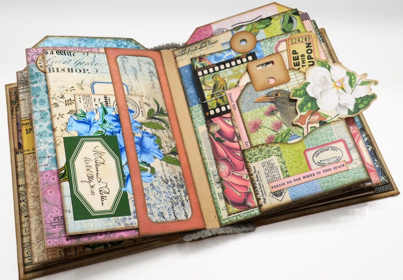 Op de afbeelding: Een handgemaakt scrapbook met een bloemenmotief. De omslag is gemaakt van bruin papier en heeft verschillende decoratieve elementen, waaronder bloemen, vogels en linten. Het scrapbook is open om verschillende pagina's te laten zien die gevuld zijn met meer decoratieve elementen en tekst.
