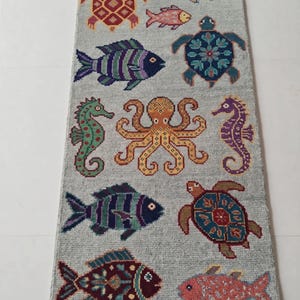 Op de afbeelding: Een rechthoekig lichtgrijs vloerkleed met een herhalend patroon van kleurrijke zeedieren, waaronder schildpadden, diverse vissen, een centrale octopus en zeepaardjes. Het kleed heeft een getextureerd oppervlak en korte witte franjes aan de uiteinden.