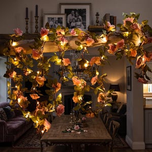 May include: A decorative garland featuring artificial pink roses and green leaves intertwined with warm white string lights. The floral vine is draped across a wooden beam, with individual roses and foliage illuminated by small, glowing LED bulbs.