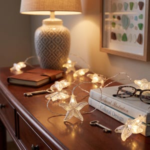 May include: A string of LED lights featuring clear, textured starfish-shaped covers. Each starfish contains a small, warm-toned light bulb. The lights are connected by a thin, flexible silver wire, creating a glowing decorative strand.