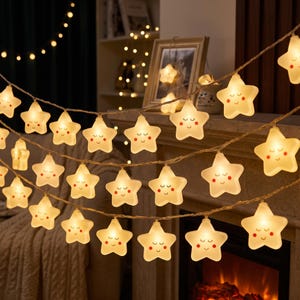 May include: A string of LED fairy lights featuring multiple star-shaped covers. Each star is off-white with a printed face showing closed eyes, a small smile, and pink cheeks. The lights emit a warm glow, connected by a thin, flexible wire.