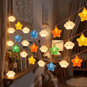 May include: A string of LED fairy lights featuring alternating star and cloud shapes. Each piece has a printed smiling face with closed eyes. The lights are illuminated in various colors including yellow, orange, blue, green, and white.
