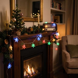 May include: A string of LED lights featuring translucent star-shaped covers in various colors including blue, green, red, orange, and yellow. The lights are draped across a wooden fireplace mantel decorated with a small Christmas tree and greenery.