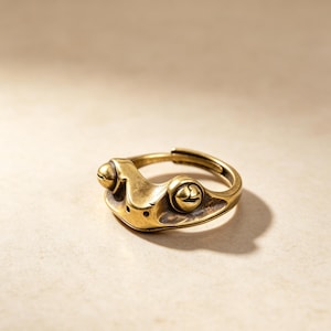 Puede incluir: Un anillo de metal en tono latón con un diseño de rana esculpida. El anillo tiene un acabado pulido con áreas oscurecidas en relieve para resaltar los ojos y los rasgos faciales de la rana. La banda está abierta en la parte posterior.