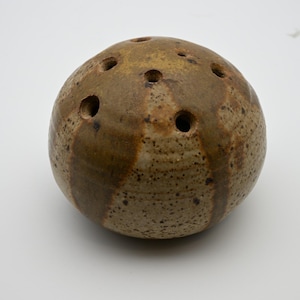 May include: A spherical ceramic flower frog with a speckled tan and brown glaze. The object features a rounded top surface with several circular holes distributed across the dome, designed for arranging stems in a floral display.