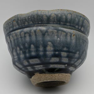 May include: A ceramic bowl with a thick, uneven rim and a textured surface. The exterior features a deep blue glaze that drips downward over a lighter, mottled gray base. The bottom of the bowl is unglazed, revealing a coarse, tan-colored clay.