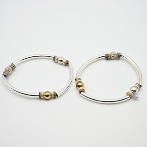 May include: Two silver-toned bangle bracelets featuring alternating accents of smooth gold-toned spheres, textured silver-toned rings, and spherical beads encrusted with small clear crystals. The bracelets have a polished finish and a circular shape.