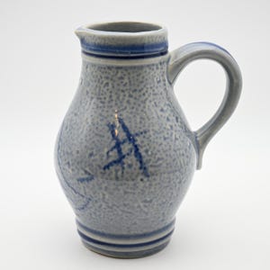 May include: A stoneware pitcher featuring a speckled light gray glaze with dark blue accents around the rim, base, and handle. The body of the pitcher displays a hand-painted dark blue mark resembling a stylized letter or symbol.