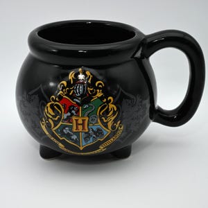 May include: A black ceramic mug shaped like a cauldron with three small feet and a large handle. The front features a colorful crest with a central letter H, surrounded by a lion, snake, badger, and eagle, with the text Draco Dormiens Nunquam Titillandus.