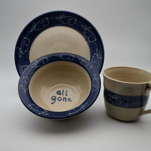Peut inclure: Un ensemble de vaisselle en céramique comprenant une assiette, un bol et une tasse. Chaque pièce présente une base crème mouchetée avec un bord bleu foncé orné de dessins au trait blancs de lapins. L'intérieur du bol affiche le texte all gone.
