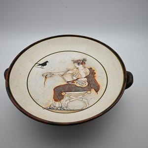 May include: A shallow ceramic bowl with two side handles, featuring a cream-colored interior decorated with a dark brown illustration of a seated figure playing a lyre, accompanied by a small black bird perched nearby.