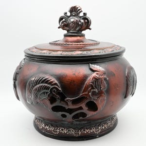 May include: A round, dark copper-colored lidded jar featuring a relief design of a rooster on the exterior. The lid is topped with an ornate, decorative finial, and the base is accented with a detailed, repeating scrollwork pattern.