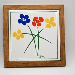 May include: A square ceramic tile framed in light brown wood featuring a white surface with a graphic illustration of four stylized flowers in blue, red, and yellow with green stems. The text "HIMARK 1983" and the signature "Sultera" are visible.