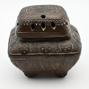 May include: A dark brown, square-shaped incense burner with a removable lid. The exterior features intricate embossed geometric patterns, circular motifs, and triangular carvings. The lid includes small ventilation holes, and the base rests on four short, sturdy feet.