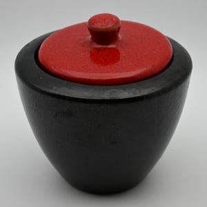 May include: A small, round, black container with a smooth, matte finish and a slightly tapered base. It features a vibrant, glossy red lid with a visible crackle glaze texture and a small, rounded knob handle centered on top of the lid.