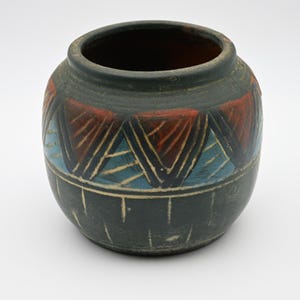 May include: A round, dark gray ceramic pot featuring a wide opening and a matte finish. The exterior is decorated with a band of alternating red and light blue triangular patterns, accented by etched lines and a series of vertical markings at the base.