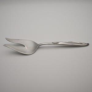 Op de afbeelding: Een zilverkleurige metalen serveervork met een brede kop met twee tanden en een gebogen handvat. Het handvat is voorzien van een in reliëf aangebracht bloem- of bladachtig decoratiepatroon bij de hals en de punt, met een gladde afwerking.