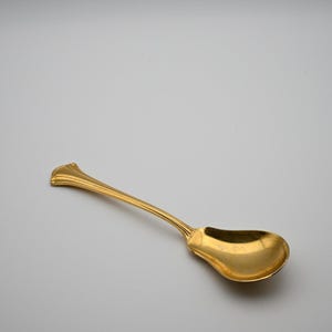 May include: A gold-colored metal spoon featuring a wide, rounded bowl and a slender, tapered handle. The handle terminates in a flared, fan-like decorative end with subtle vertical ridges, while the neck displays a small, engraved pattern.