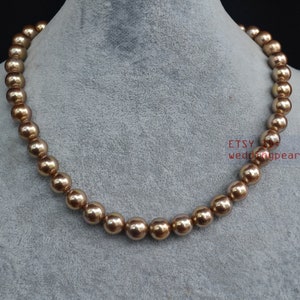 Puede incluir: Una hebra de perlas de color marrón dorado sobre un fondo gris. Las perlas son redondas y lisas, y están enhebradas en un hilo fino y transparente.