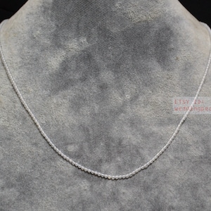 Puede incluir: Un delicado collar de perlas blancas sobre un fondo gris. El collar está hecho de pequeñas perlas redondas que están enhebradas en un hilo delgado. El collar mide aproximadamente 46 cm de largo.