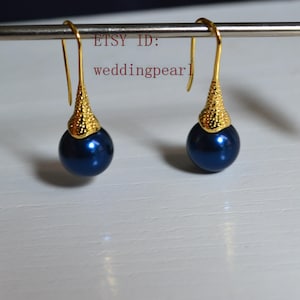 May include: A pair of navy blue pearl earrings with gold-colored textured tops and gold-colored hooks. The earrings are hanging from a silver bar. The text "ETSY ID: weddingpearl" is visible.