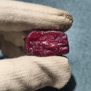 Puede incluir: Una piedra preciosa en bruto de color rojo intenso con una superficie rugosa y texturizada de bordes irregulares. La piedra muestra un tono carmesí saturado con sutiles variaciones en el reflejo de la luz sobre su estructura cristalina desigual.