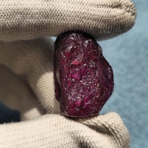 Peut inclure: Une pierre précieuse rubis brute d'un rouge profond avec une surface texturée et inégale. La pierre présente une teinte cramoisie sombre et saturée avec des facettes cristallines naturelles et des inclusions visibles sur sa forme organique irrégulière.