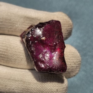 May include: A raw, deep red gemstone with a polished, irregular surface texture. The stone displays translucent qualities with dark crimson and purple hues throughout its crystalline structure, showcasing natural facets and a glossy, reflective finish.