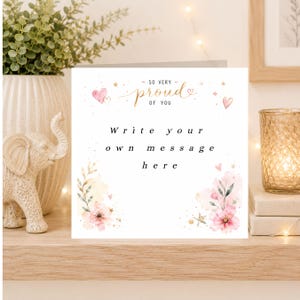 May include: A square greeting card with a white background featuring the gold text SO VERY PROUD OF YOU at the top and the black text Write your own message here in the centre. The design includes pink watercolour flowers and gold stars.