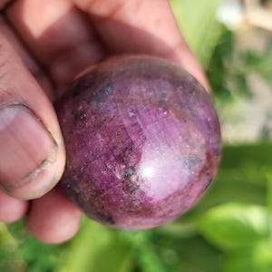 Peut inclure: Une pierre précieuse sphérique polie présentant une palette de couleurs magenta profond et violet foncé avec des inclusions cristallines visibles et des textures de surface naturelles sur toute la forme arrondie, avec une finition brillante.