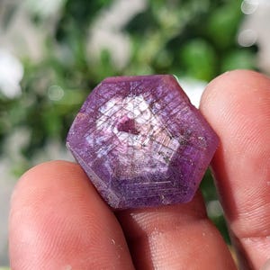 May include: A hexagonal, translucent gemstone with a deep magenta and purple hue. The surface displays a distinct geometric pattern of intersecting lines and internal inclusions, with a polished, flat top reflecting light in a bright, central spot.