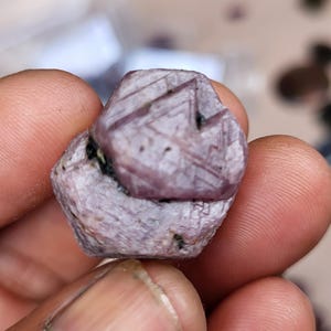 Könnte beinhalten: Ein roher, sechseckiger Rubinkristall mit einem staubigen, blassvioletten und rosafarbenen Farbton. Die Oberfläche weist natürliche geometrische Streifen, geätzte Dreiecksmuster und kleine dunkle Einschlüsse auf, die eine raue Textur zeigen.
