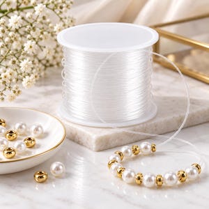May include: A spool of clear, flexible elastic cord for jewellery making. A portion of the cord is threaded with alternating white faux pearl beads and small, polished gold-tone metal spacer beads, forming a circular loop on the surface.