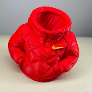 May include: A bright red, quilted puffer jacket-shaped container with a high collar and a central zipper line. The garment features a yellow Nike swoosh logo on the upper left chest area. The design mimics a miniature, puffy winter coat.