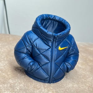 May include: A miniature dark blue puffer jacket figurine featuring a quilted diamond pattern, a high collar, and a small yellow Nike swoosh logo on the upper left chest. The jacket is designed with a full-length front zipper detail.