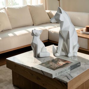 May include: Two white geometric animal sculptures, one larger and one smaller, featuring faceted surfaces and sharp angles. The sculptures are positioned on a white marble surface next to a book with the text "FLANSUNT" visible on the spine.