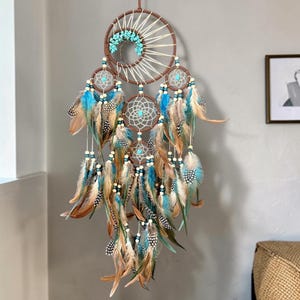 May include: A decorative dreamcatcher featuring a large upper hoop with a tree-like design made of turquoise stones, connected to three smaller lower hoops with intricate web patterns. It is adorned with numerous blue and brown feathers.