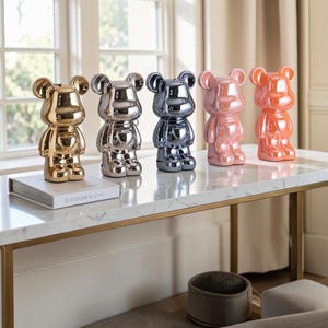 May include: Five bear-shaped figurines with a glossy, reflective finish arranged in a row. The colours from left to right are gold, silver, dark grey, light pink, and orange. Each figurine features rounded ears, a smooth body, and a heart shape on the chest.