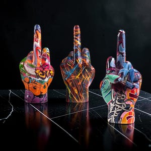 May include: Three hand sculptures displaying the middle finger gesture, each featuring a unique, vibrant graffiti-style paint finish with colourful patterns, swirling lines, and abstract shapes covering the entire surface of the upright hand figures.