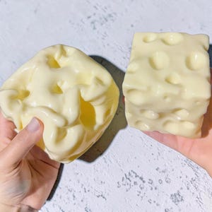 Könnte beinhalten: Zwei cremeweiße, glänzende, käseförmige Objekte mit unregelmäßigen Löchern. Ein Objekt hat eine abgerundete, organische Form mit einer glatten, welligen Oberfläche, während das andere eine klare Würfelform mit mehreren kreisförmigen Vertiefungen ist.