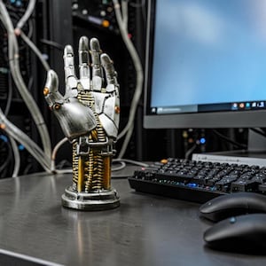 May include: A robotic hand sculpture featuring a metallic silver finish with exposed gold-colored mechanical wiring and circuitry details. The hand is mounted on a circular base, showcasing intricate segmented plating and small orange accents.
