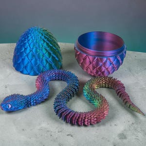 Peut inclure: Une figurine de serpent articulée et flexible avec des écailles irisées aux reflets bleus, violets et verts. À côté du serpent se trouvent deux moitiés d'un œuf texturé assorti, l'une bleue et l'autre violette, avec une finition métallique.