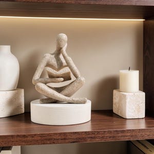 Può includere: Una statuetta umana astratta, color beige e maculata, seduta a gambe incrociate mentre tiene in mano un libro aperto. La figura appoggia la testa su una mano ed è montata su una base circolare bianca e liscia, con una finitura in pietra ruvida.
