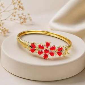 May include: A gold-toned hinged bangle bracelet featuring a central floral motif composed of vibrant red pear-shaped stones and small clear accent crystals. The metal band has a smooth finish with ribbed detailing near the hinge mechanism.