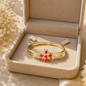 Puede incluir: Un brazalete dorado con un motivo central en forma de cruz, adornado con cuatro piedras rojas en talla pera y cristales transparentes en talla baguette. El brazalete se presenta dentro de un joyero forrado en terciopelo beige con tapa.